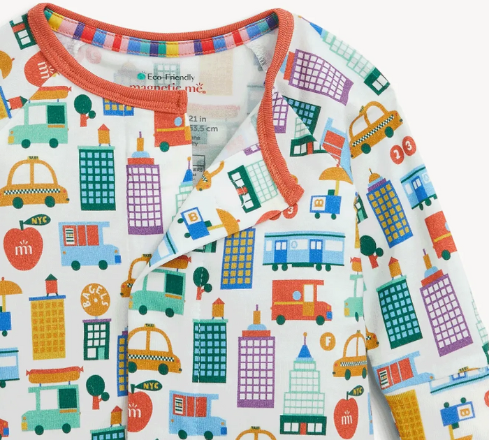 A close-up of a children's long-sleeve top with a colorful city print on a white background. The garment features orange trim along the collar and front wrap, with a pattern of yellow cabs, purple skyscrapers, green trees, and red apples.