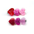 Two heart-shaped hair clips on a white background, one set with smooth glossy red, pink, and light pink hearts, and the other set with glittery red, magenta, and lavender hearts.