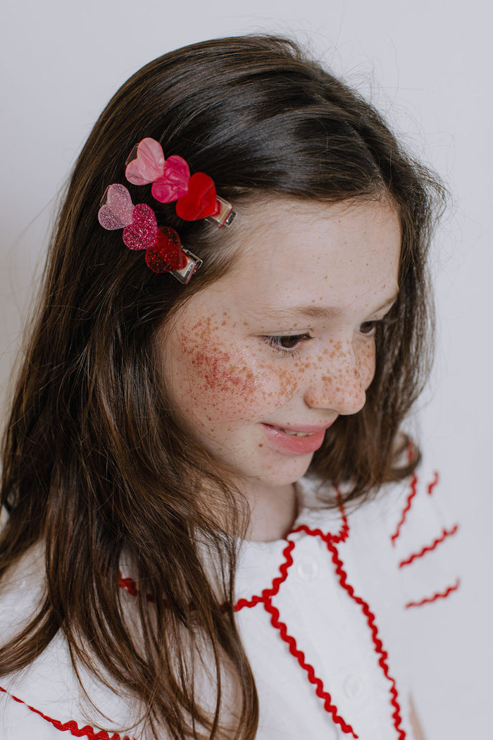 A close-up of a young girl with brown hair and freckles, wearing a cluster of heart-shaped hair clips in shades of pink, red, and purple. She is wearing a white top with red embroidered details, set against a plain light gray background.