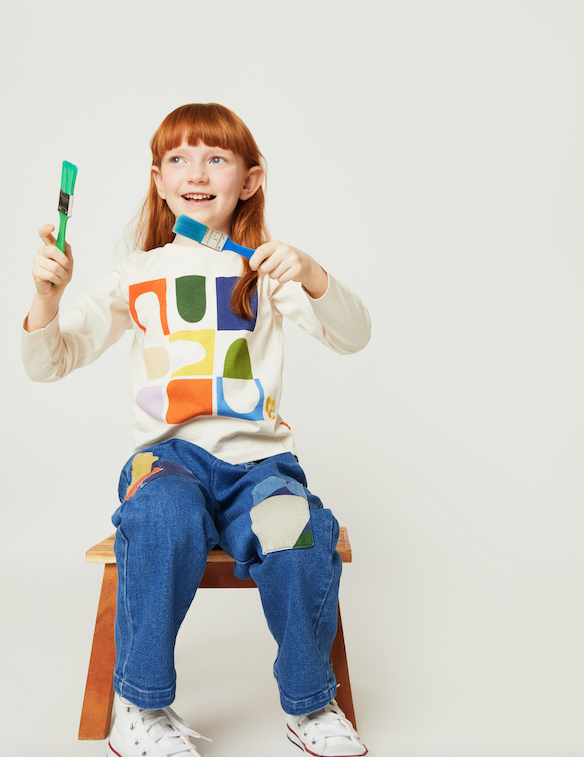 A young girl with red hair sits on a wooden stool against a light gray studio background, wearing a cream long sleeve shirt with a colorful geometric patch design, blue jeans with patch details, and white sneakers. She holds up a green paintbrush in one hand.